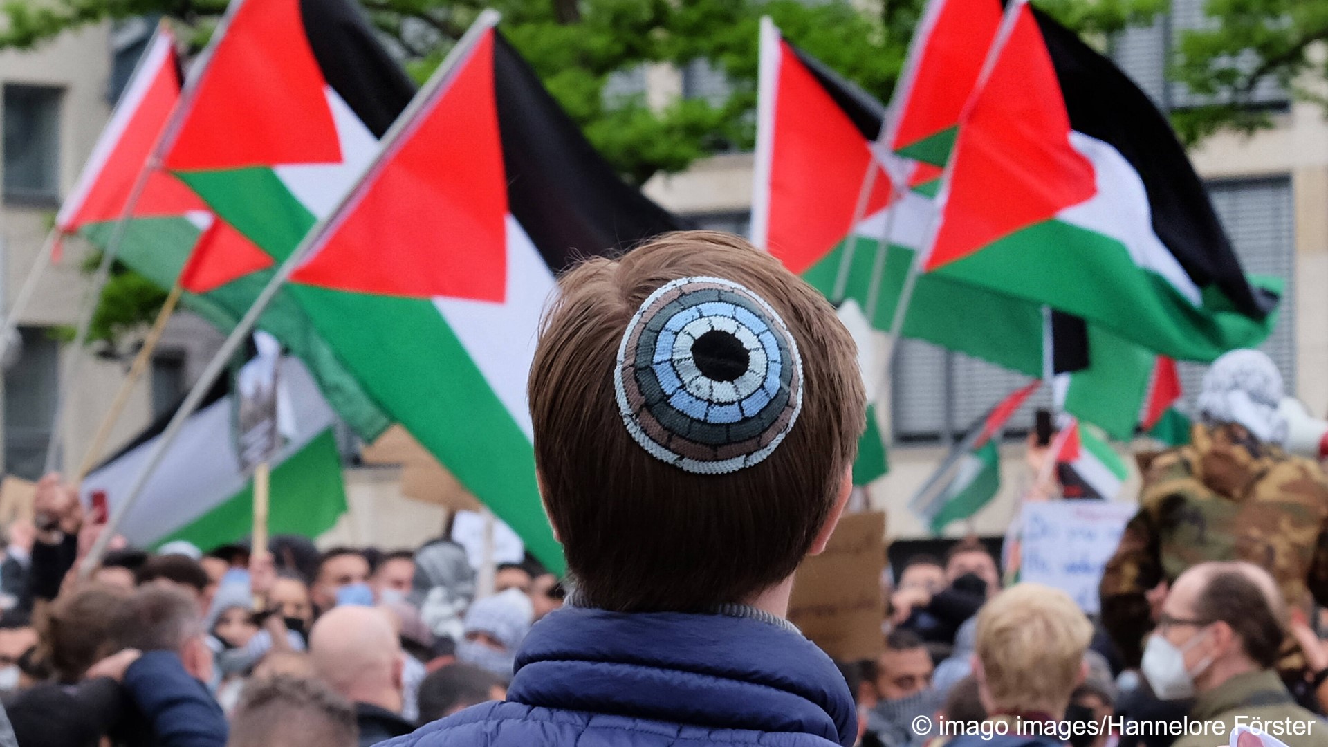 A man wearing a kippah with his back to the camera, against a sea of Palestinian flags carried by a large group of people