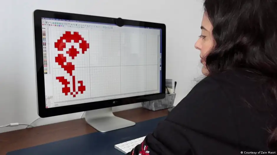 Woman sits in front of a screen displaying a cross-stitch pattern of a red flower