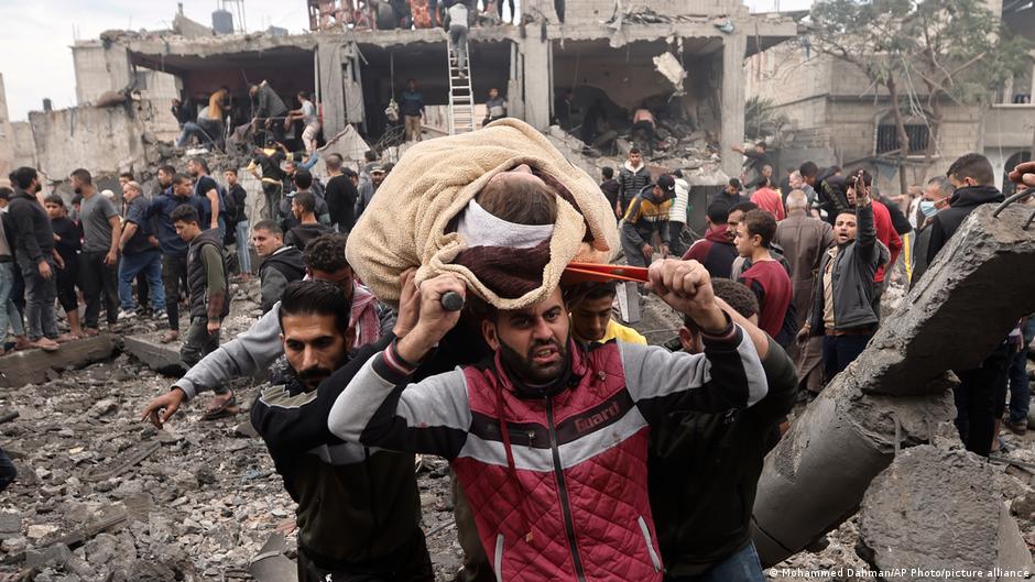 A person wrapped in a blanket is carried away from a ruined house on a stretcher