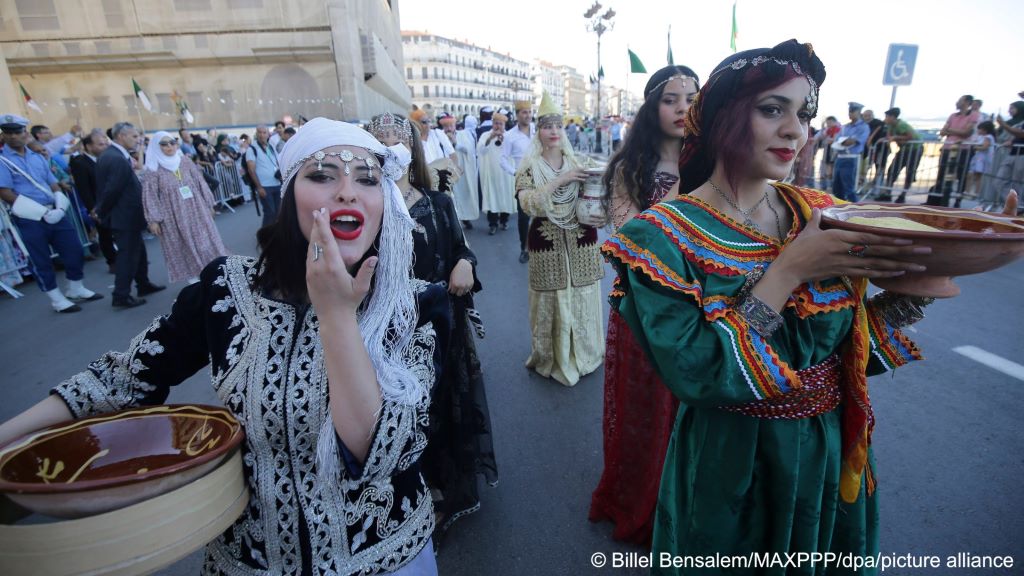 Women in traditional costumes and carrying large bowls and containers walk down the middle of a street as part of a protest on the 61st anniversary of Algerian independence while people on the footpaths and police officers look on