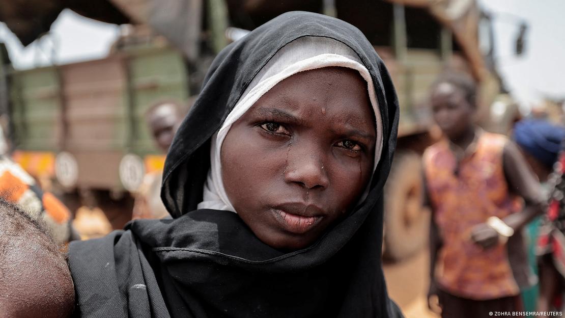 A black woman wearing black and white headscarves looks into the camera as tears run down her face