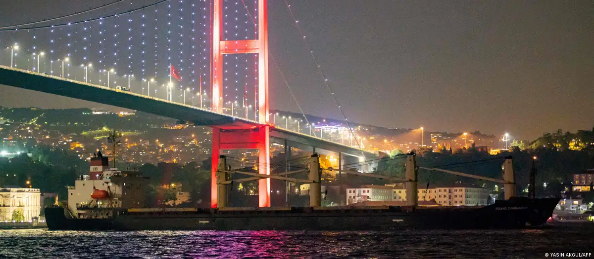 The Bosporus Bridge is illuminated in red and white at night; the lights of Istanbul can be seen in the background as a ship passes beneath the bridge