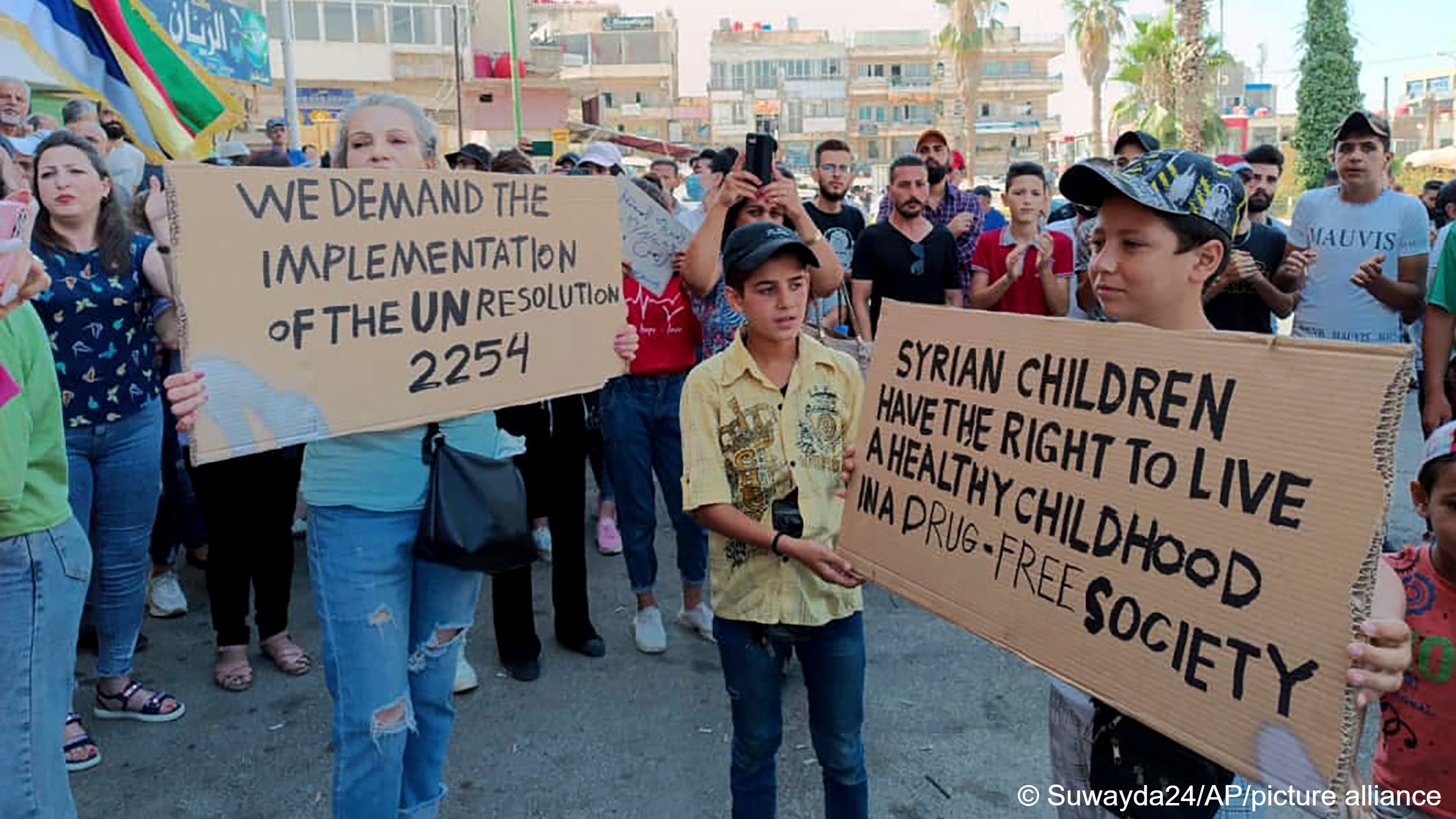 صورة من: Suwayda24/AP/picture alliance - ارتفاع الأسعار أطلق شرارة احتجاجات السويداء. A woman and children hold cardboard placards demanding the implementation of UN Resolution 2254 and a drug-free society. In the background is a crowd of people, some clapping, some holding up Druze flags, Sweida, Syria, 26 August 2023