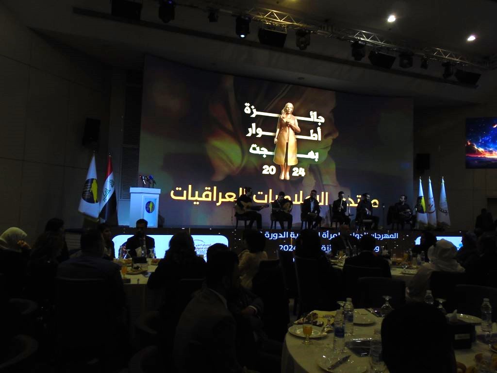 A group of musicians perform on stage in front of a screen showing the statuette of a woman in a darkened room in front of people sitting at round tables