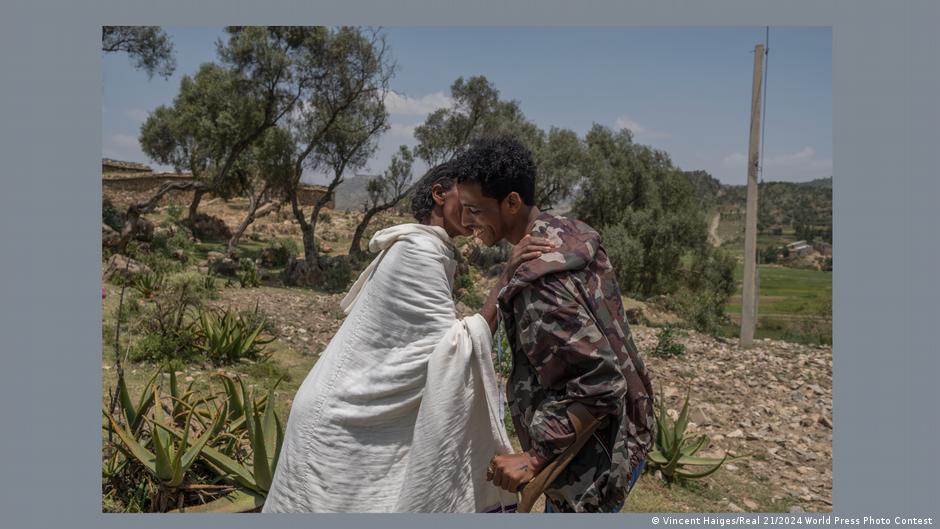 A woman dressed in a loose white garment kisses her son, who is wearing military fatigues and walking with crutches, on the cheek