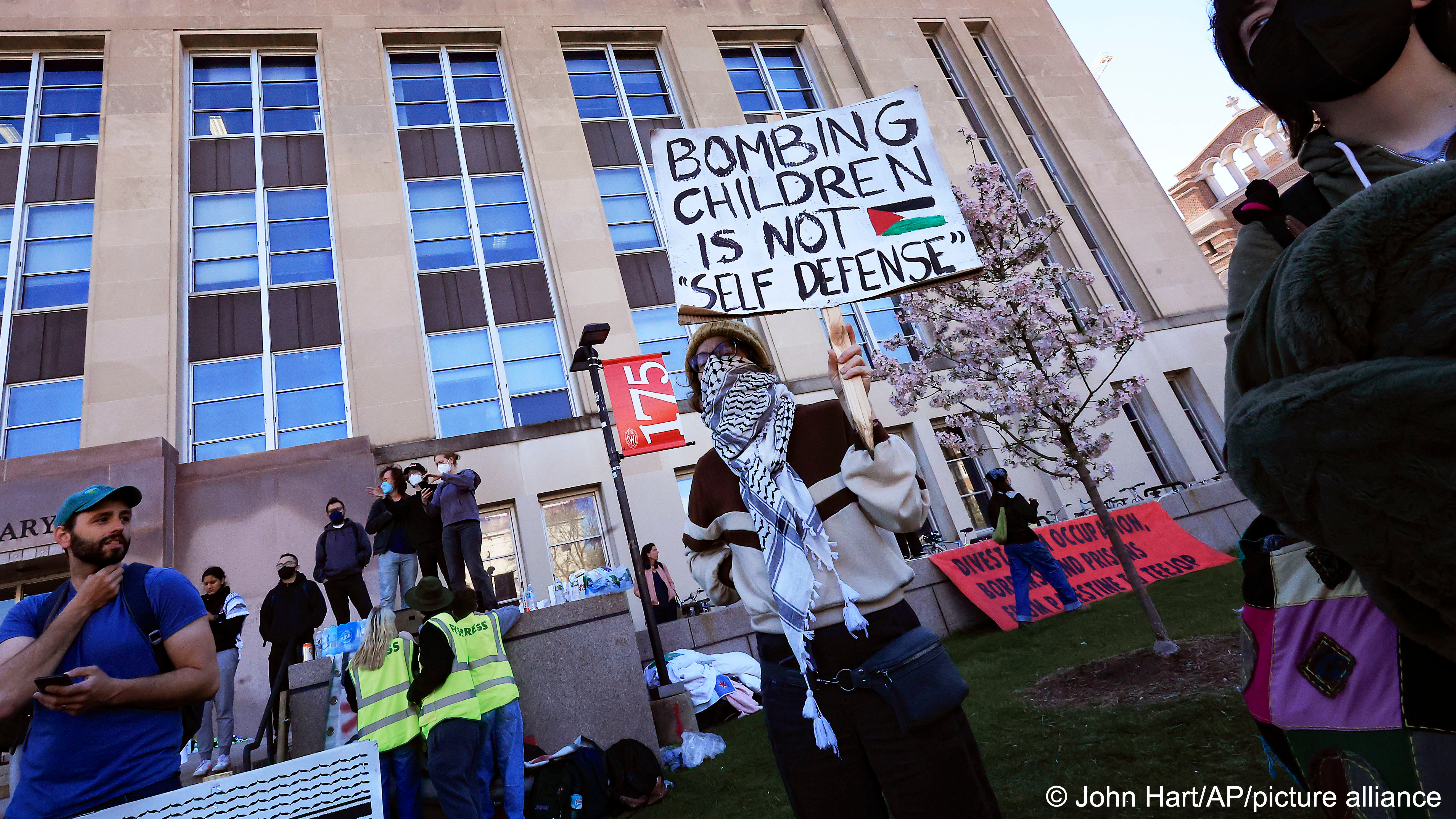 Masked demonstrators against the war in Gaza watch the dismantling of a tent encampment by law enforcement personnel on the campus of UW-Madison in Madison, Wisconsin, USA., 1 May 2024. One demonstrator holds up a placard that reads "Bombing children is not 'self defense'"