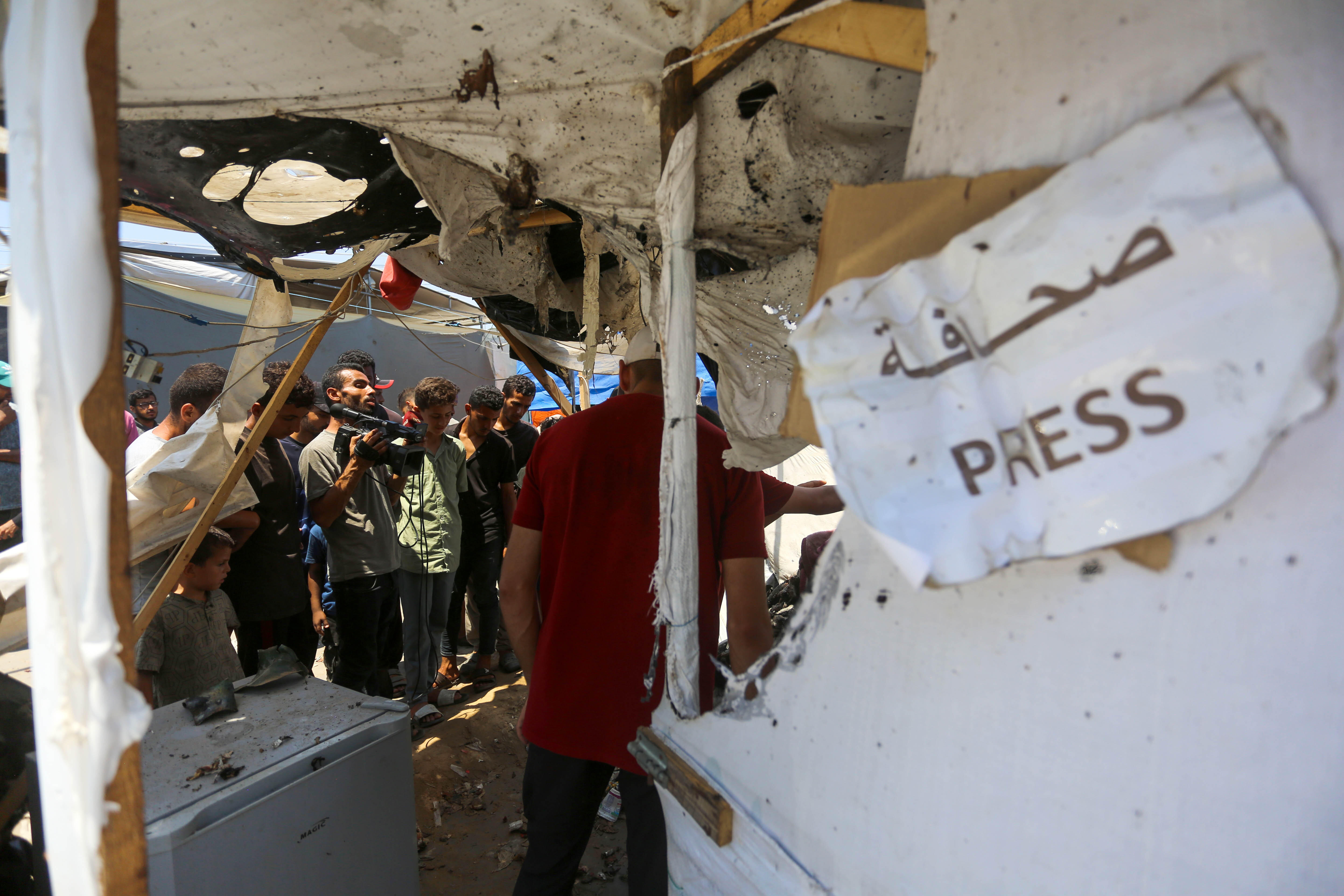 View of a destroyed tent in Gaza. In the foreground hangs a sign that reads ‘Press’ in Arabic and English. Journalists can be seen in the background.