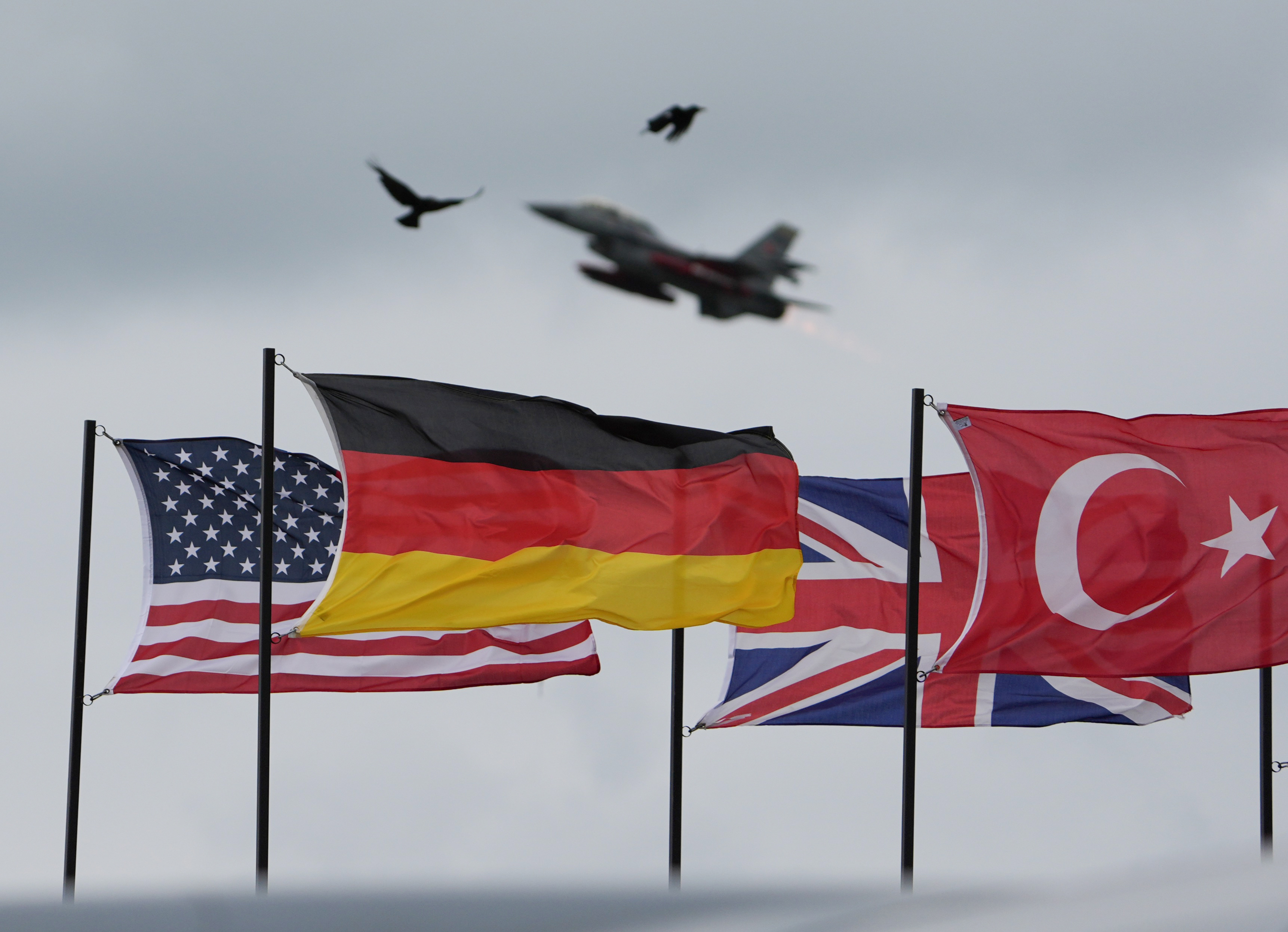 American, German, English and Turkish flags are waving in the wind. A NATO fighter jet flies above them.