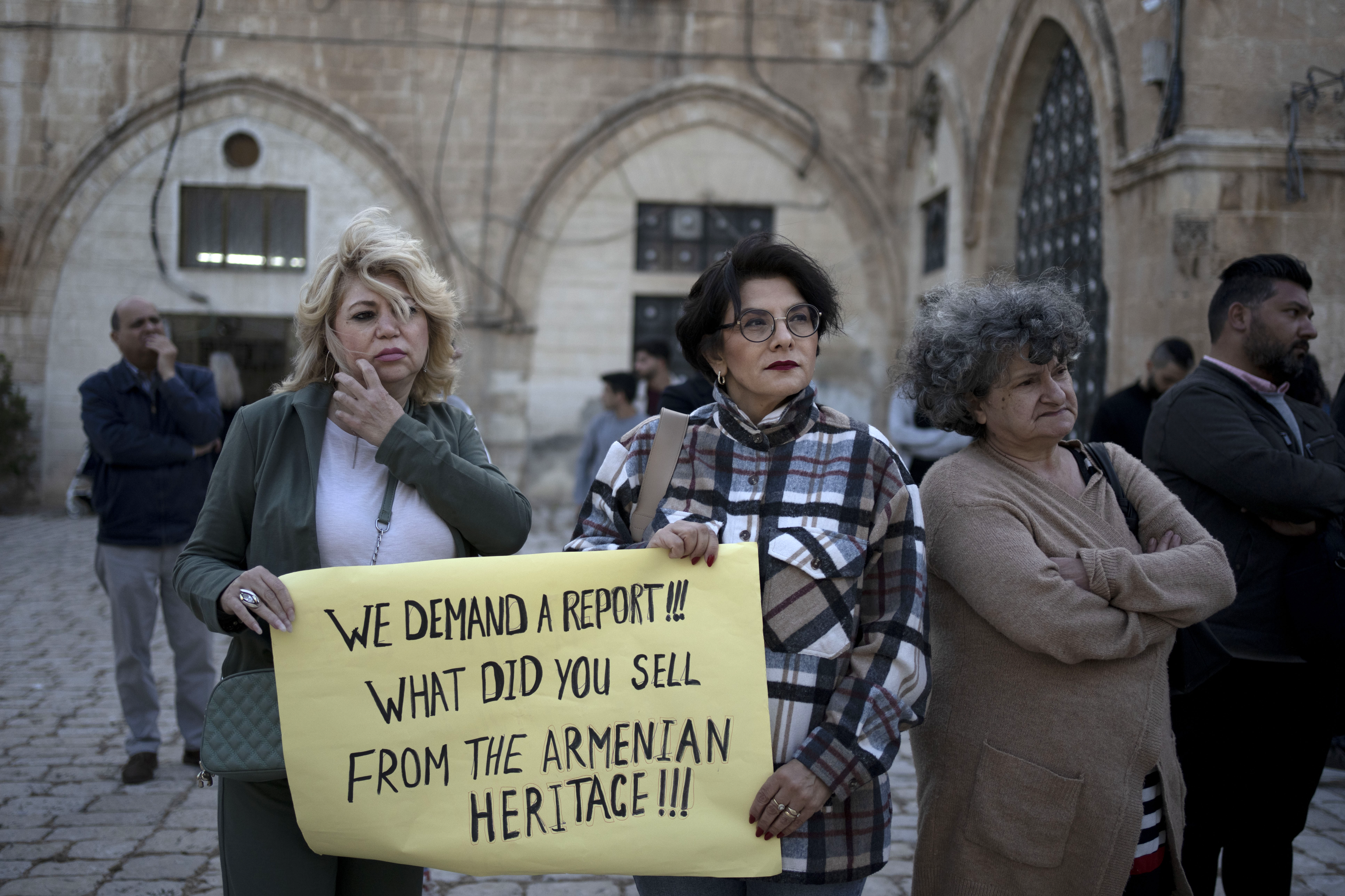 Drei Demonstrantinnen auf einem Platz in der Altstadt Jerusalems. Eine von ihnen hält ein Schild in den Händen, auf dem steht: Wir verlangen einen Bericht! Was vom armenischen Erbe habt ihr verkauft?"