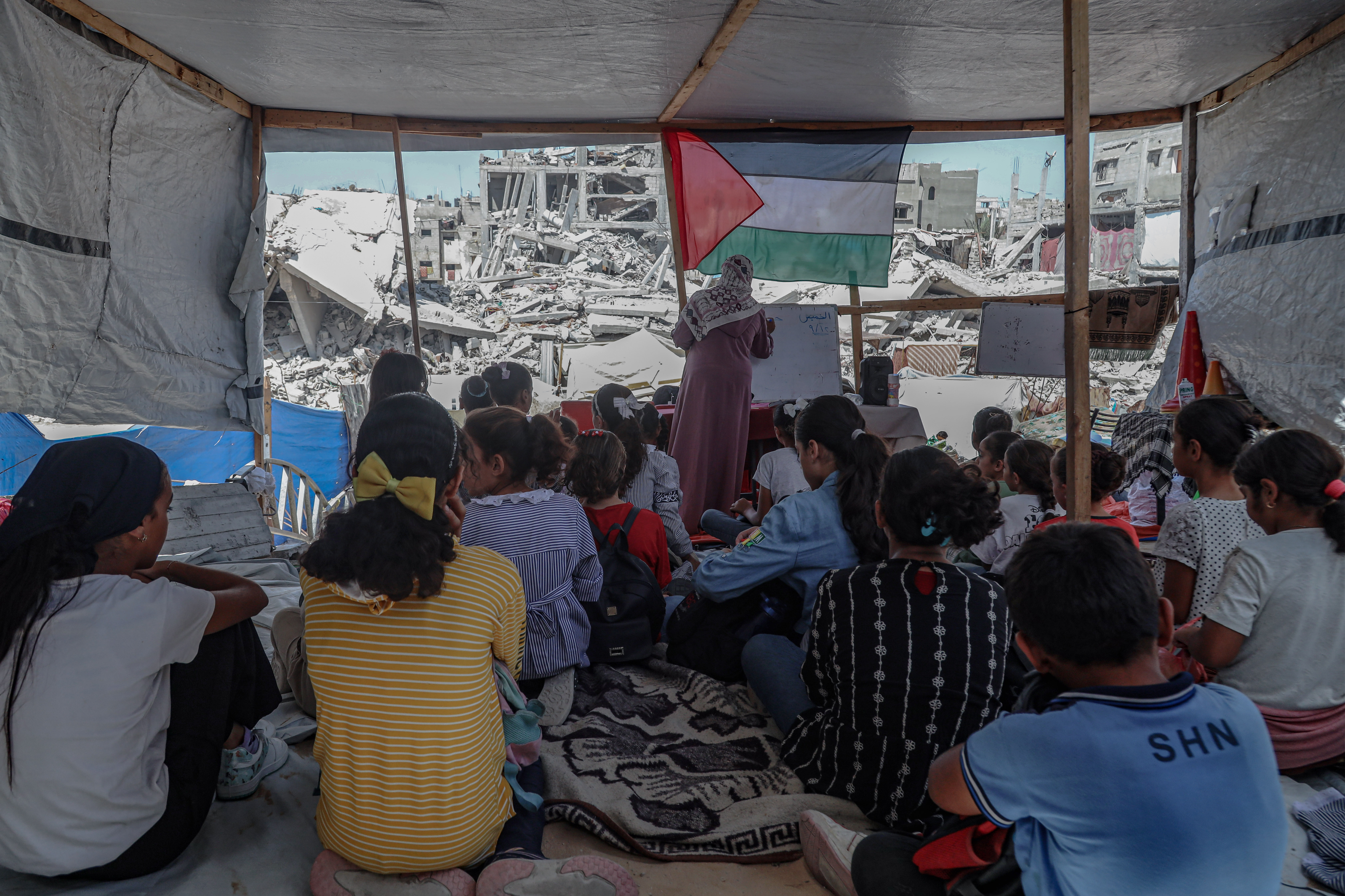 A group of children sit on the floor of a ruined house. A teacher stands at the front, behind her is a Palestinian flag and a view of a destroyed building.