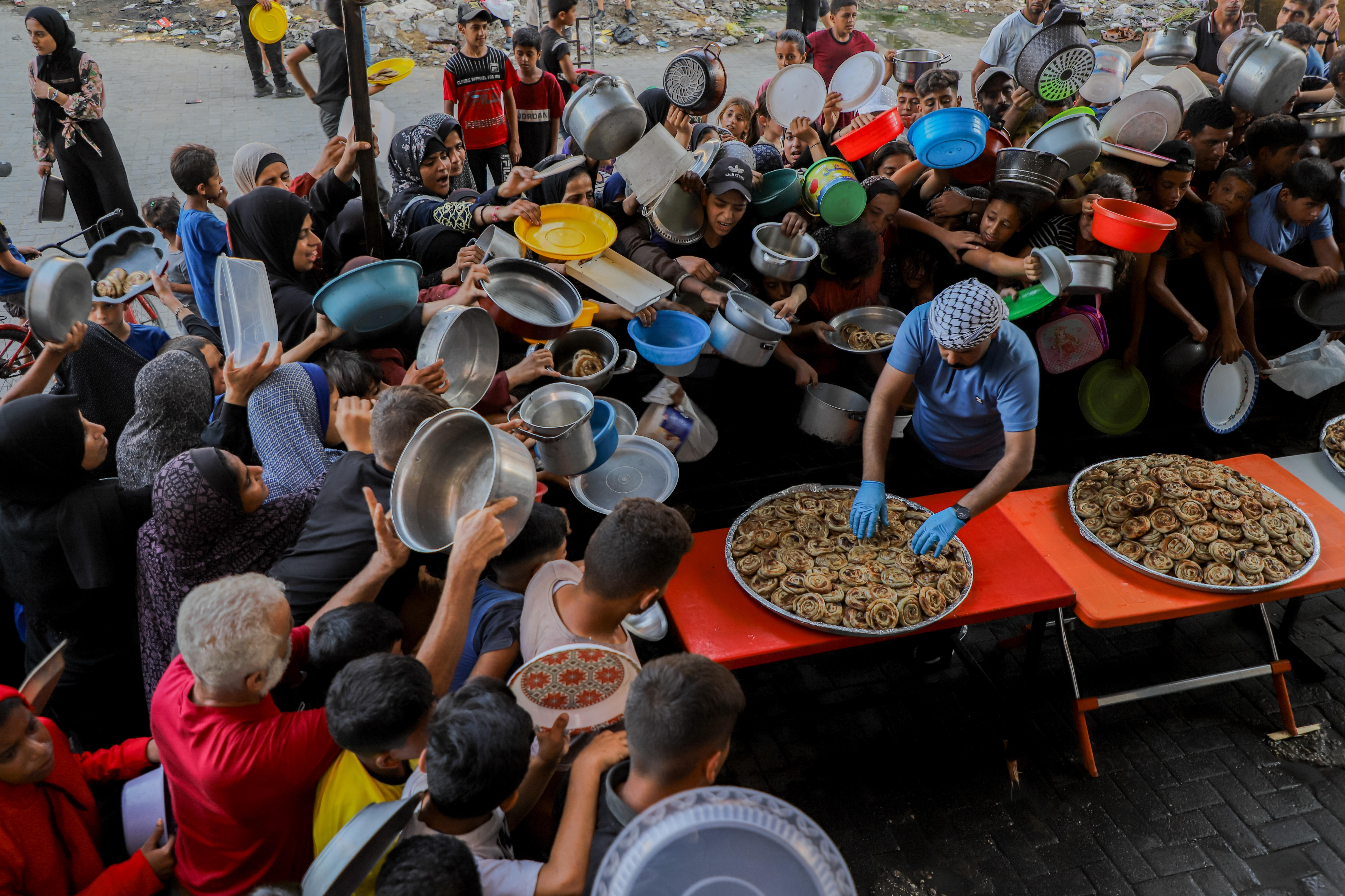 A large group of people holding empty plates, bowls and other recepticles crowd around an aid worker serving food from a large pan.