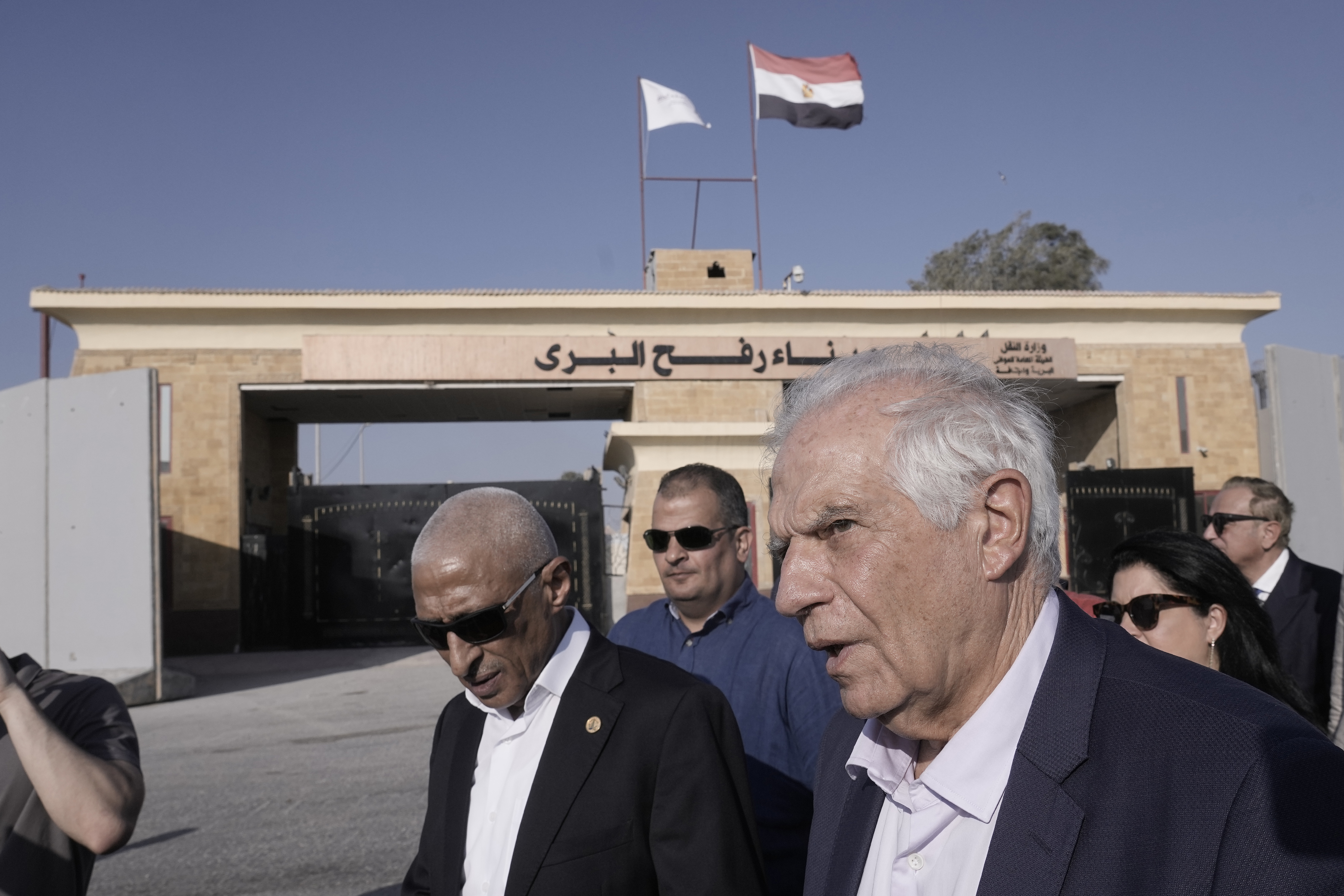 Josep Borrell walks alongside a group of people in suits. In the background is the Rafah border crossing with the flag of Egypt on top.