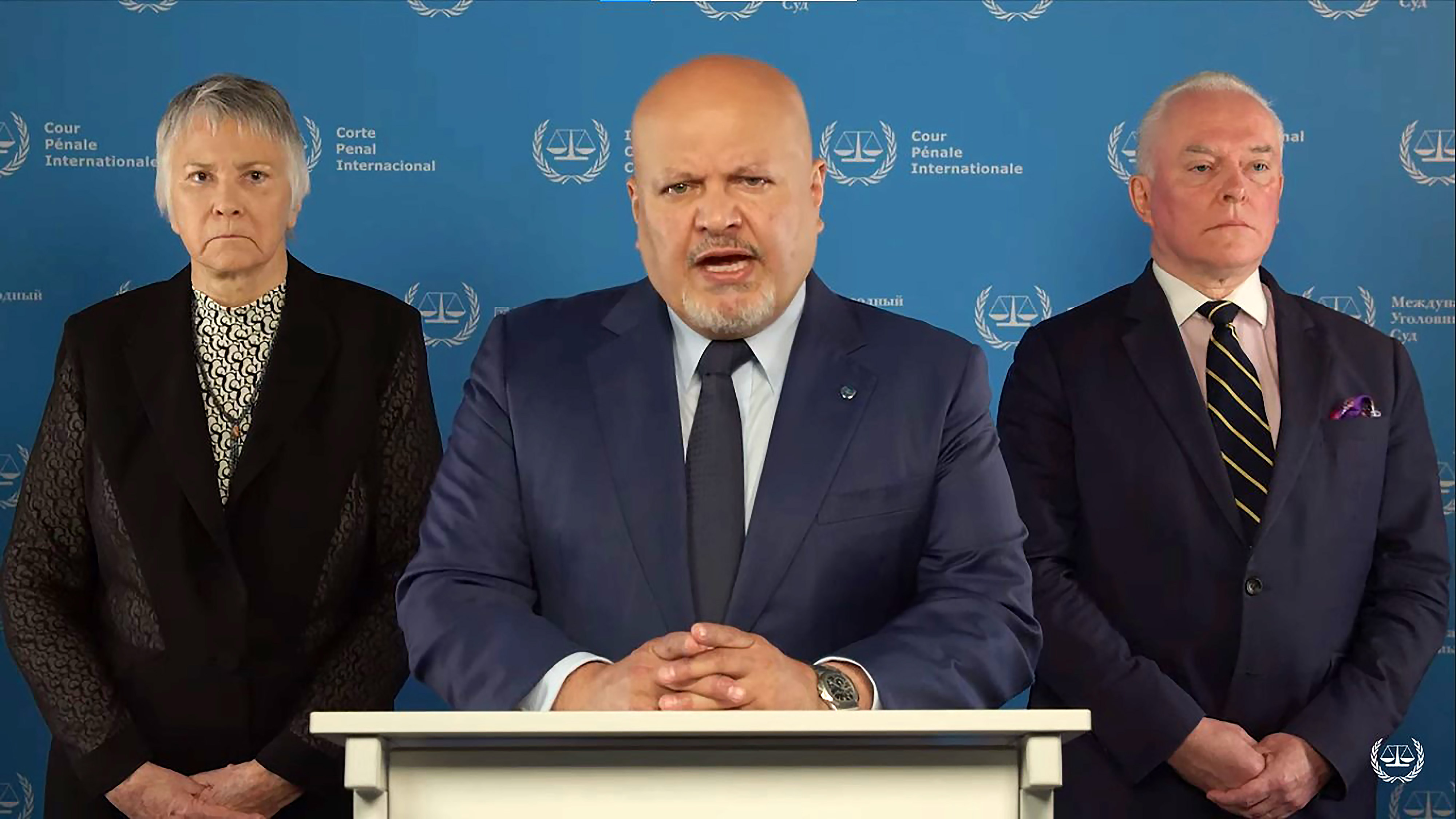 Chief prosecutor of the ICC Karim Khan and gives a speech to the camera. There is a person to his left and one to his right.