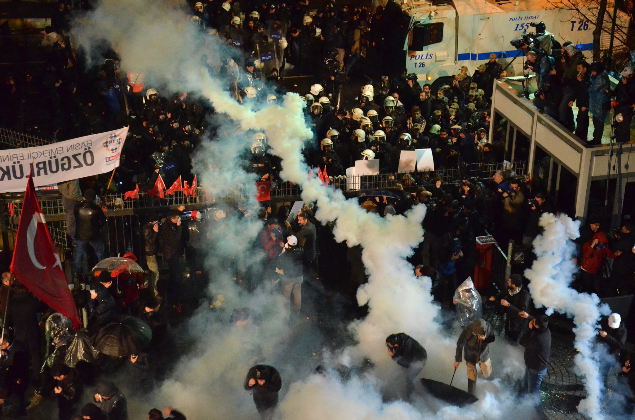 A group of police clash with a crowd of protesters. Smoke rises from among the protesters, one carries a Turkish flag on a pole.