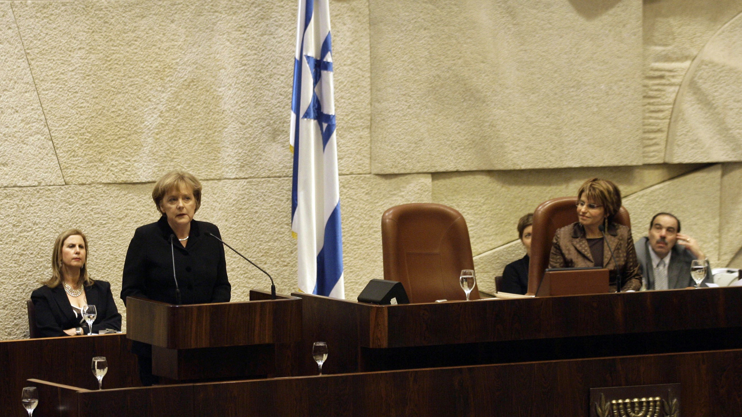 Angela merkel stands at a podium speaking into a microphone. An Israeli flag hangs in the background.