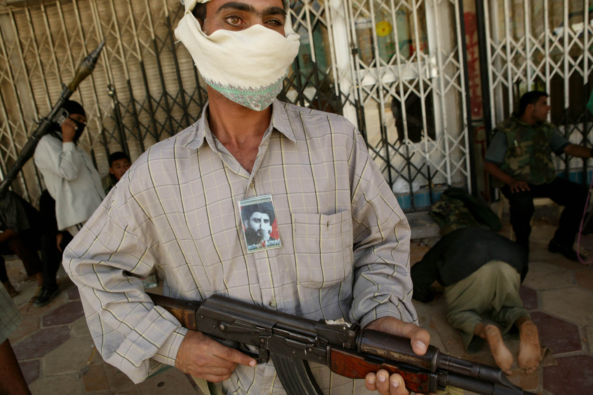 Fighter with a picture of Moqtada Sadr on his chest holds a machine gun while another fighter prays in the background (photo: Michael Kamber)