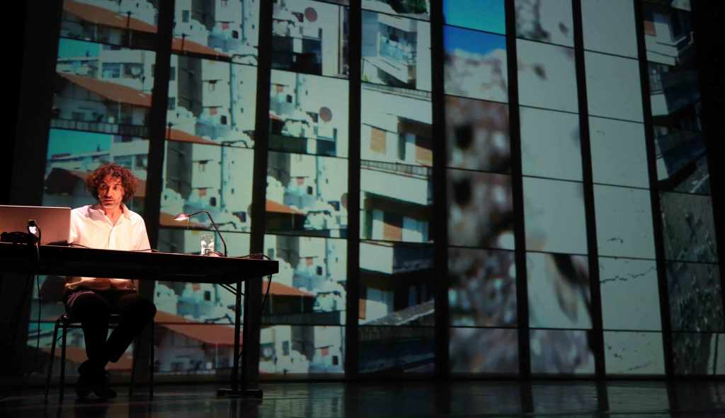 Rabih Mroué sits in front of a projection during his lecture performance "The Pixelated Revolution" (source: Kampnagel.de)