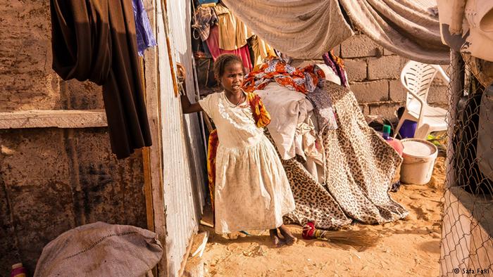 A young girl wearing a dress stands alone in a makeshift house