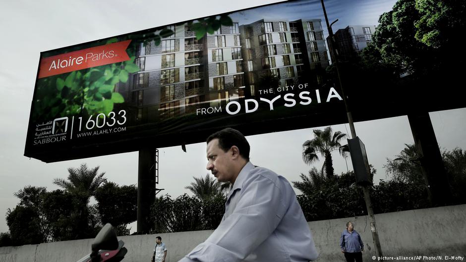 A man rides his motorbike as others wait for a bus under a billboard promoting a new residential housing compound, in Cairo, Egypt; October 2018 (photo: AP Photo/Nariman El-Mofty)