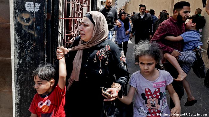 Crying woman holds hands of children, fleeing for safety; a man follows carrying a child