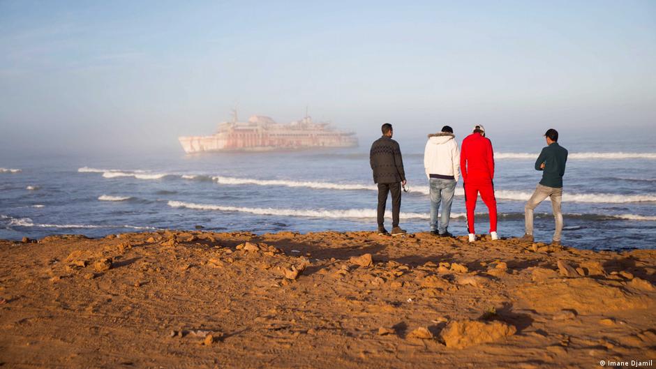 Hassan Boulahcen, 27, ein Surftrainer, Maatoug, Hossin Ofan, 34, der allgemeine Koordinator des Surfclubs Nuevas Olas, und Oussama Segari, 26, der als Schatzmeister des Clubs und Manager des Coffeeshops arbeitet, blicken vor dem Armas-Schiffswrack auf das Meer. Foto: Imane Djamil