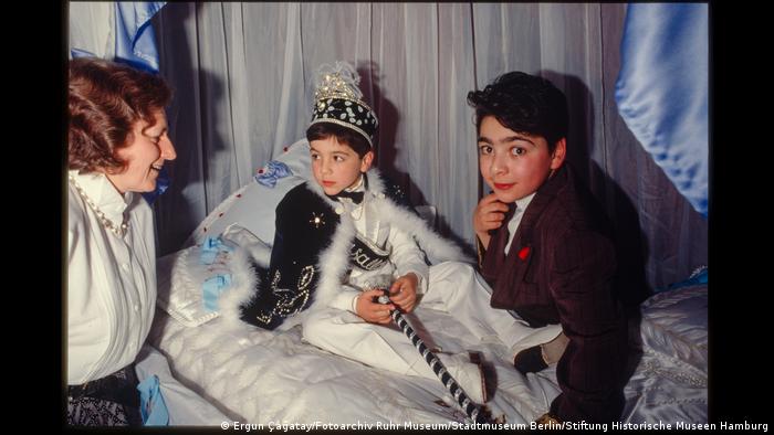 A boy in festive clothes at his circumcision party, with another boy and a woman next to him