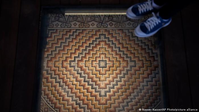 Feet stand on a see-through platform, beneath which can be seen an illuminated geometric mosaic