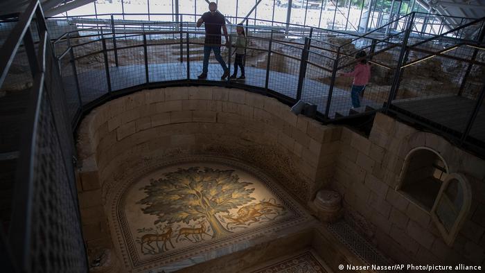 A person walks on a walkway past an intricate mosaic depicting a tree and animals