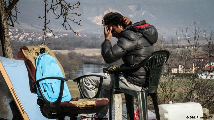 A man sits on a plastic chair, a river or lake and mountains can be seen in the background