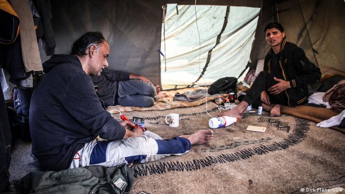  An older and a younger man sit on a blanket in a tent, between them lies another person whose upper body is not visible