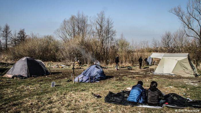 Tents in a meadow, people in winter clothing sit in front of them, behind them bushes and trees can be seen that are not in leaf