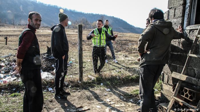 An unshaven man looks into the camera, three others stand in front of a fence and talk to two men in yellow tabards