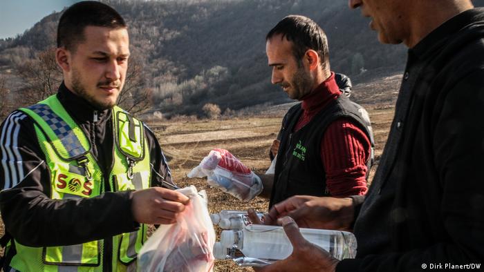 A man with a beard and a yellow tabard hands plastic bottles and filled plastic bags to other men