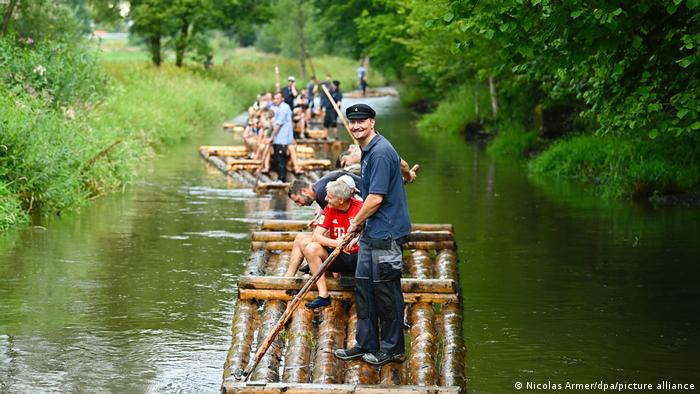 A raftsman steers a raft with a wooden batten, on which other people are sitting. Other rafts can be seen in the background