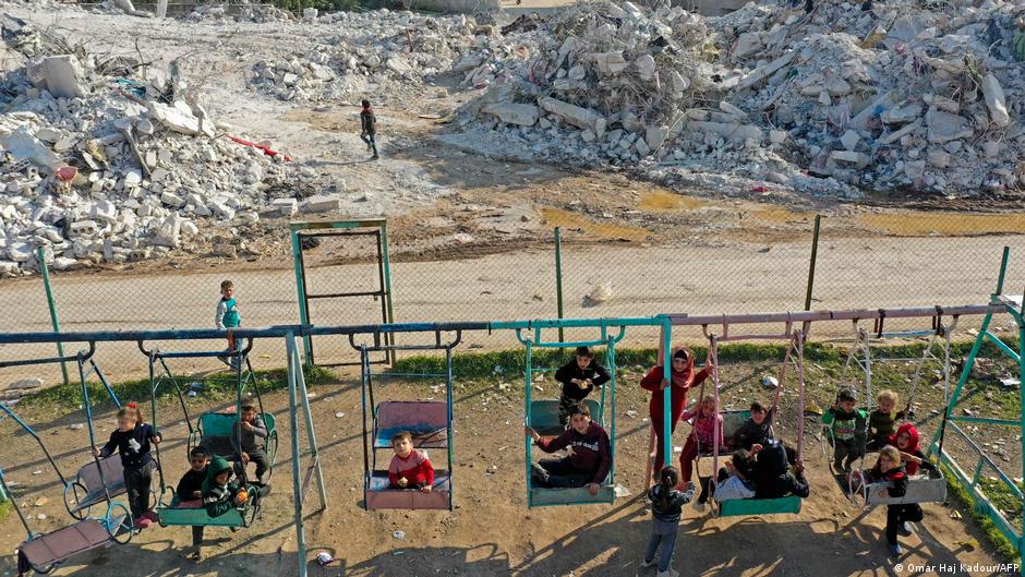 Kinder spielen auf Schaukeln, im Hintergrund eine Straße aus Trümmern; Foto: AFP