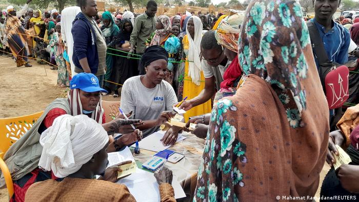 People sitting at an outdoor table receiving certificates, in the background several people are waiting in a long queue.