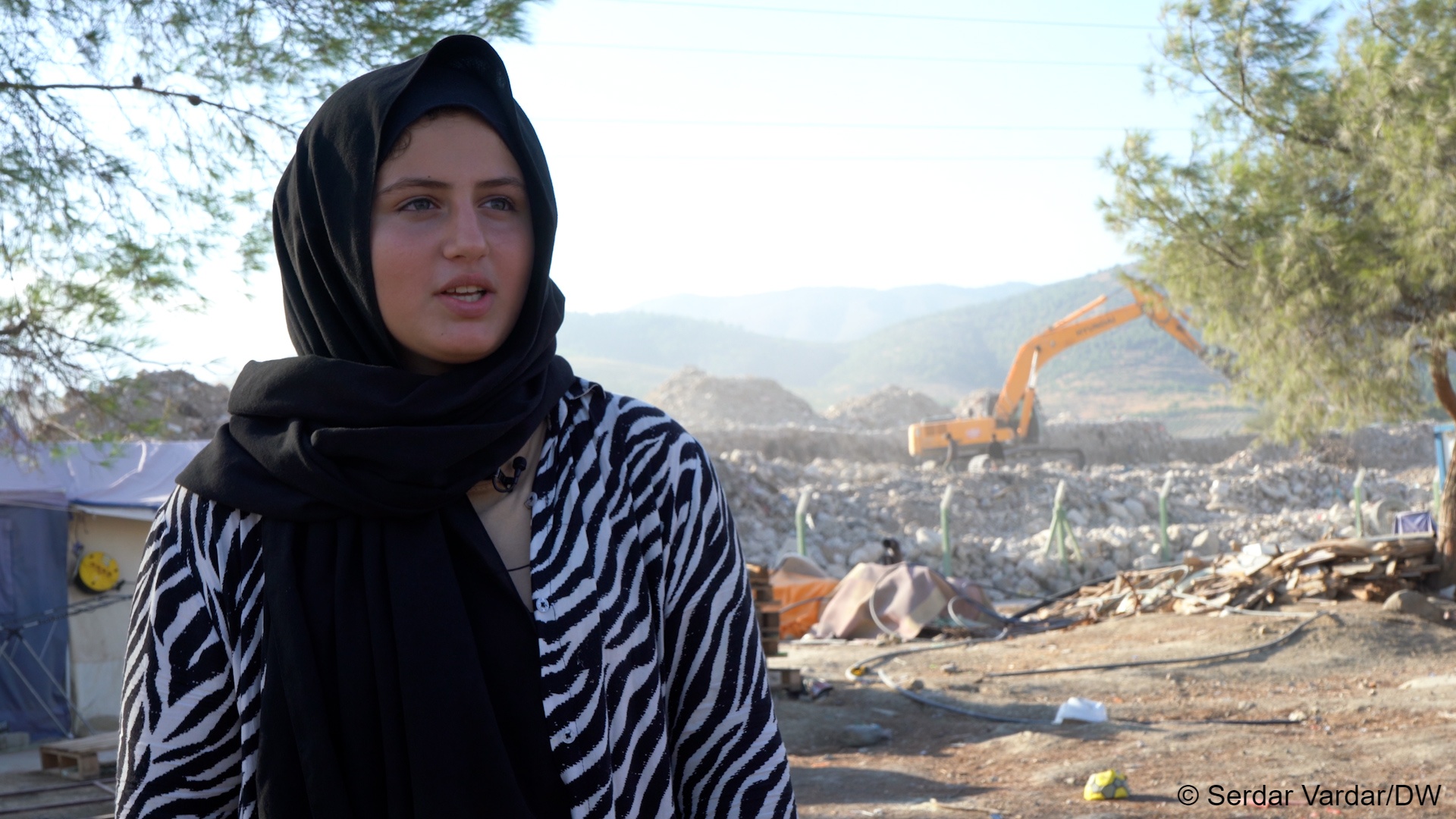 Limar Yunusoglu speaks, while a yellow excavator clears rubble in the background (image: Serdar Vardar/DW)