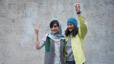 Two women stand arm in arm in front of a wall and show the victory sign. 