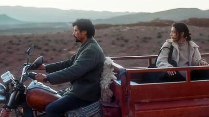 A man sits astride a motorbike while a woman sits in the trailer behind, against a background of hilly dry terrain 