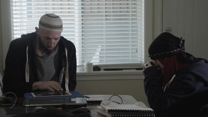 A man and a woman sit at a desk, a window shielded by a blind in the background