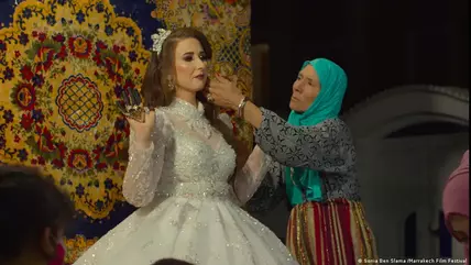 A woman in a blue headscarf puts the finishing touches to a bride's make-up; the bride looks unhappy