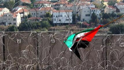 A Palestinian flag is caught in barbed wire near the Israeli West Bank barrier; houses can be seen in the background