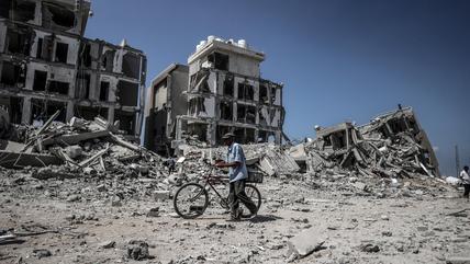 A man pushes a bicycle through rubble. In the background are three destroyed buildings.