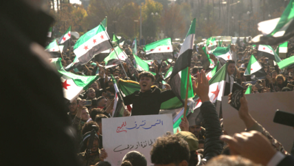 A crowd of people celebrating, many Syrian revolutionary flags are being waved.