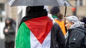 One person stands with their back to the camera, carrying an umbrella and with a Palestinian flag around their shoulders.