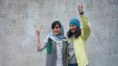 Two women stand arm in arm in front of a wall and show the victory sign. 