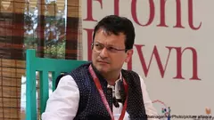 Indian art critic Ranjit Hoskote sits on a turquoise chair, a striped curtain and a white wall bearing large red letters behind him