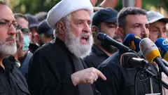 Naim Qassem wearing a white turban speaks into a microphone at a podium surrounded by men. His finger is pointing to the ground.