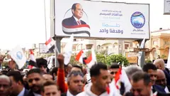 A crowd of people holding Egyptian flags. A poster with the face of President Sisi in the background.