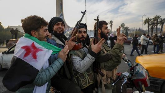 A group of young men carrying guns, one has a Syrian revolutionary flag wrapped around his shoulders.